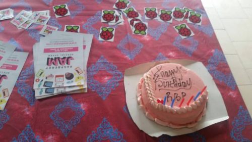 A round pink-iced cake decorated with the words "Happy Birthday RBP" and six candles, on a table beside player Pi stickers, player Jam stickers and player Jam fliers