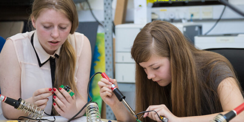 Two girls solder physical 控制早射土方法 components in a workshop.