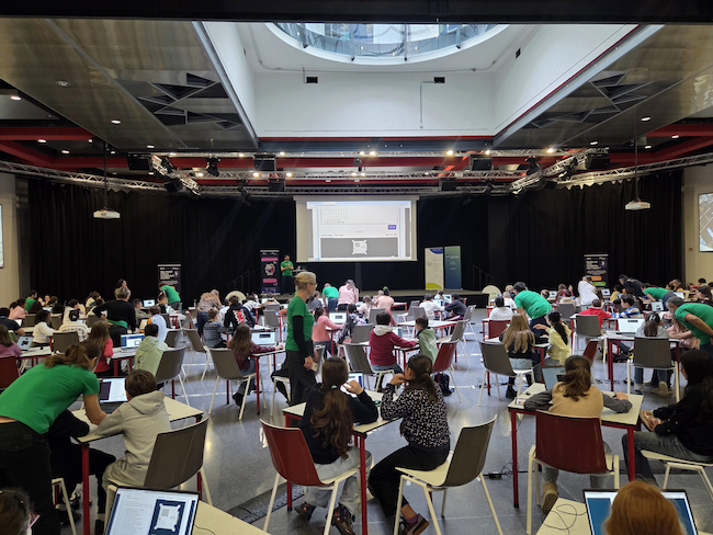 Young people coding Mission Zero at tables in a large events space.
