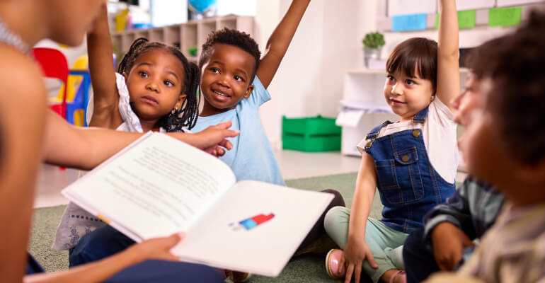 Group of four 丽柜美束 age students gathered around a teacher who is reading a picture book. Three students are raising their hands.