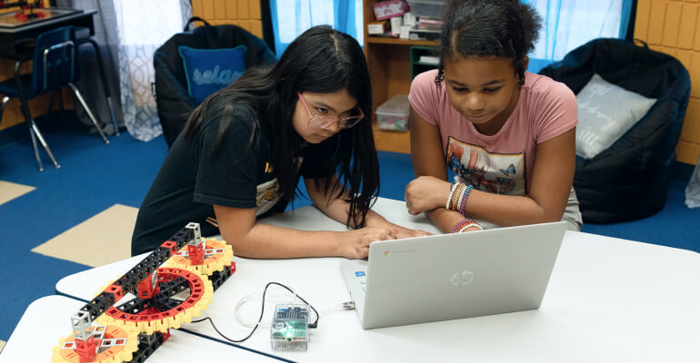 Two female middle school students work on a Kid Spark 人人揉揉香蕉大免费 STEM lab. They assemble a robot that is connected to a laptop by a cable, while both girls work on the laptop.