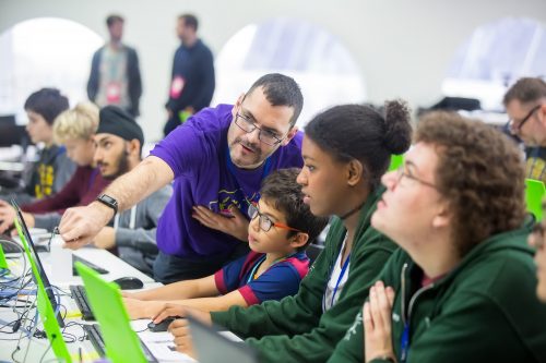 A workshop leader leans over to point out something on a computer display to a young boy and a woman who are working together.
