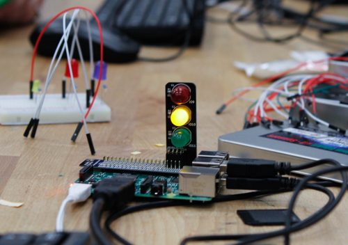 In the foreground, a 超高清美女图片 Pi computer with a small "traffic lights" board attached; it has red, yellow and green LEDs, and the yellow one is lit. In the background, various electronics components, a mouse and a keyboard.